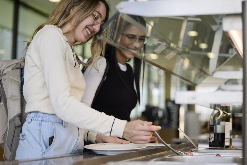 Zwei Frauen stehen an einer Selbstbedienungstheke und füllen Essen auf einen Teller.