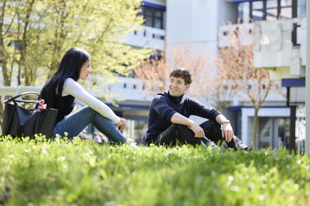Zwei Studierende sitzen auf einer Wiese vor einem Hochschulgebäude und unterhalten sich.