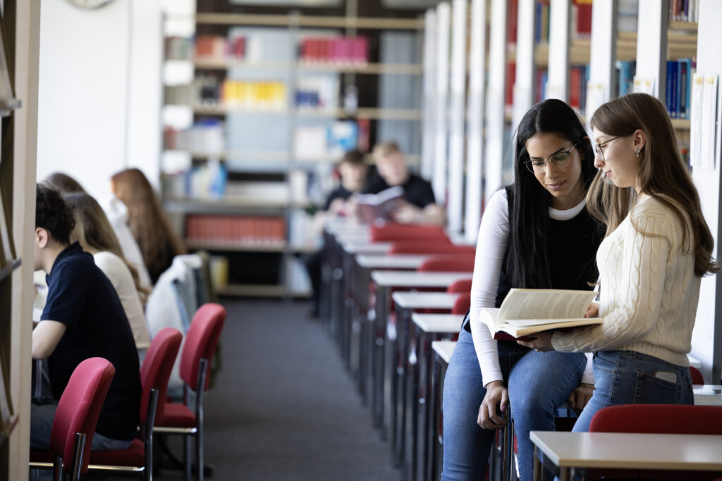 Zwei Studentinnen sitzen in einer Bibliothek und lesen gemeinsam in einem aufgeschlagenen Buch.