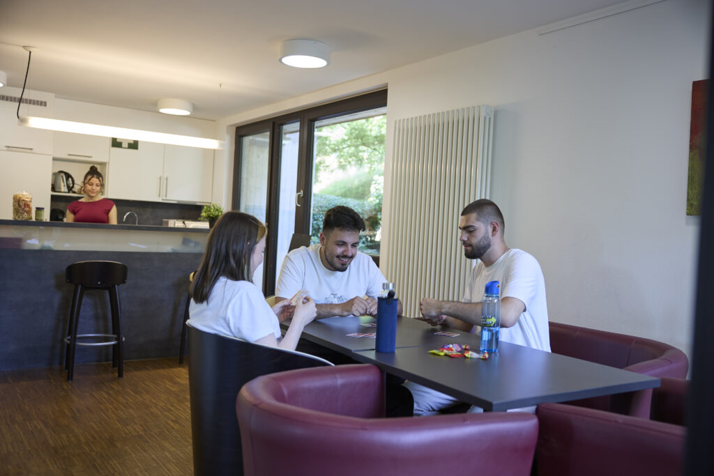 Drei Studierende sitzen gemeinsam an einem Tisch in einem Aufenthaltsraum, im Hintergrund steht eine Person in der Küche.