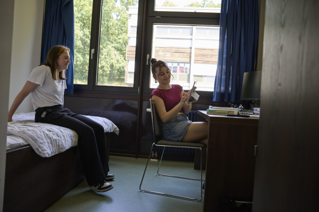 Zwei Studentinnen in einem Wohnheimzimmer, eine sitzt auf dem Bett, die andere am Schreibtisch mit einem Tablet.