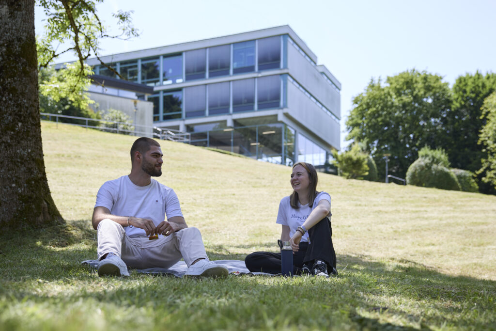 Zwei Studierende sitzen auf einer Wiese vor einem Hochschulgebäude und unterhalten sich.