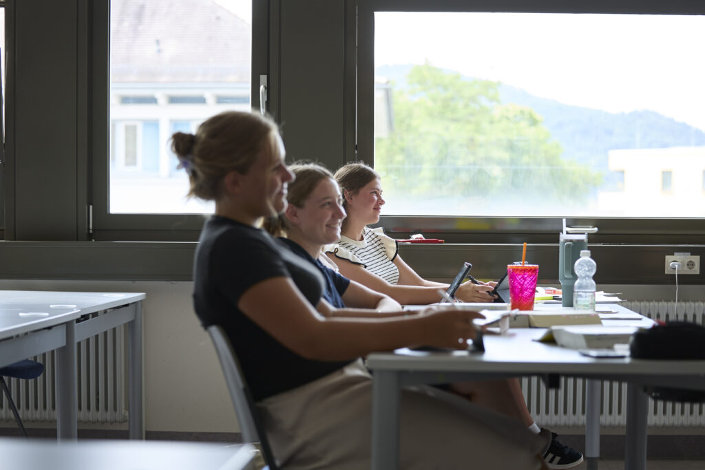 Drei Studentinnen sitzen im Seminarraum am Tisch und hören aufmerksam zu.